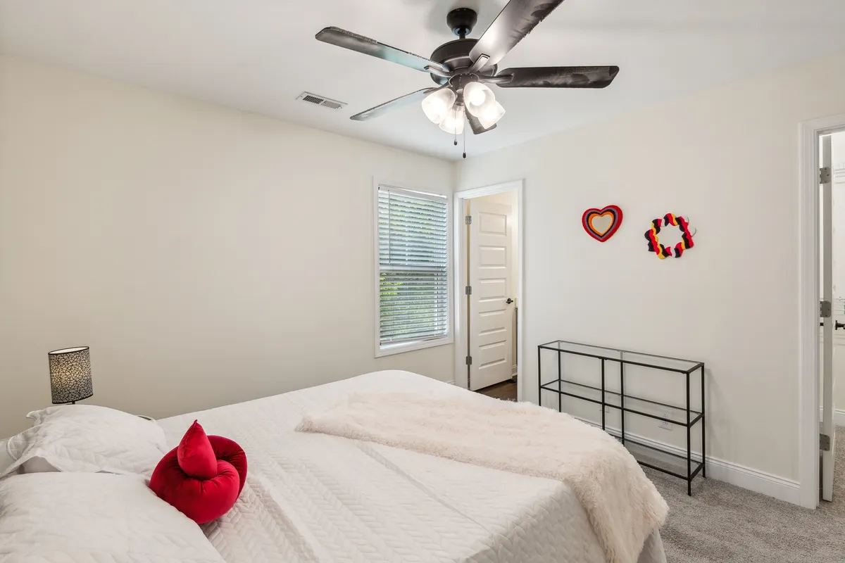 A well-appointed bedroom featuring a bed adorned with decorative pillows, a ceiling fan for added comfort, and cheerful wall decorations. The muted tones and layered textures create a soothing, restful environment.