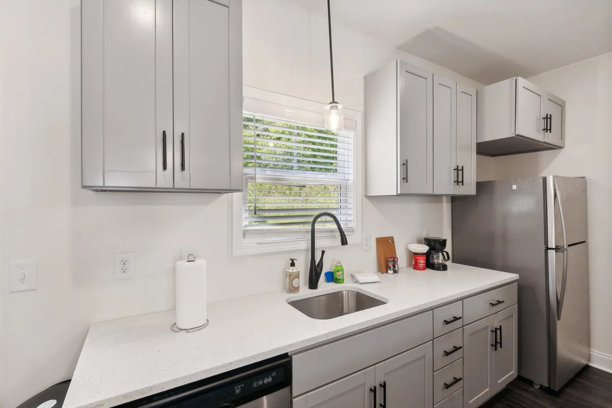 This image features a bright, functional kitchen with a spacious countertop, modern stainless-steel refrigerator, ample cabinetry, and a kitchen sink at the forefront. The mood is inviting, with hints of ongoing meal preparation and family activities. The open sightlines suggest a seamless connection to adjacent living spaces, reinforcing the kitchen’s role as a social and culinary hub.