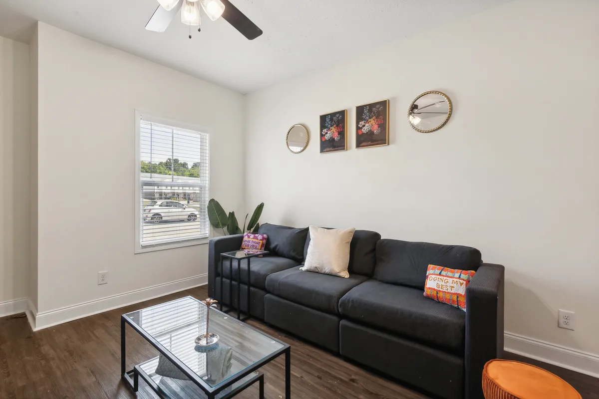 The image showcases the inviting living area of a spacious apartment, featuring a plush sofa adorned with decorative pillows, a stylish coffee table, tasteful wall art, and natural light streaming through a large window. The space exudes comfort, perfect for unwinding or sharing moments with friends and family.