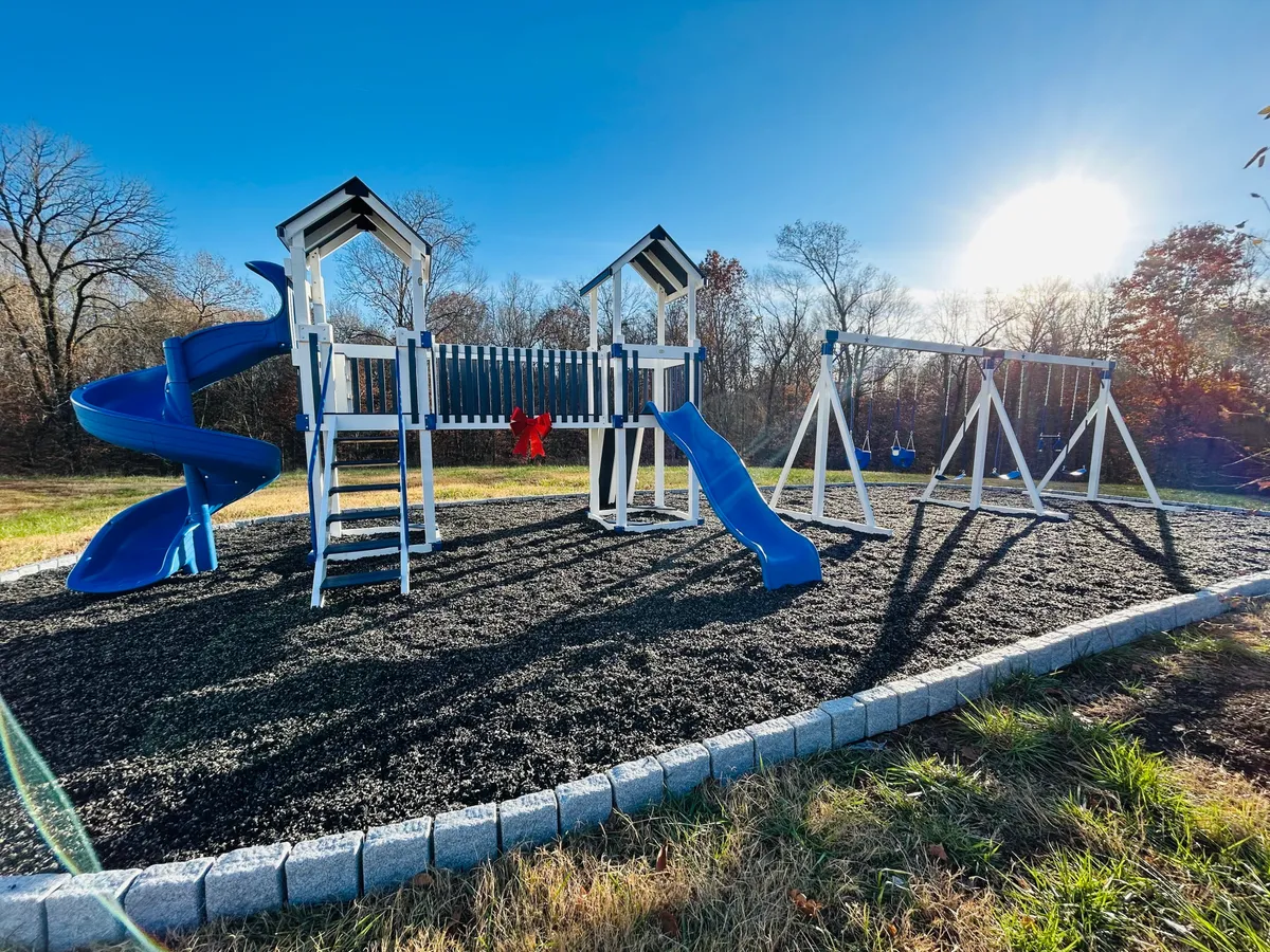This image features a cheerful playground with a blue slide and swings, surrounded by green space. Children are seen playing, while families gather nearby for picnics and socializing. The overall mood is vibrant and welcoming, emphasizing the playful outdoor amenities available for families.