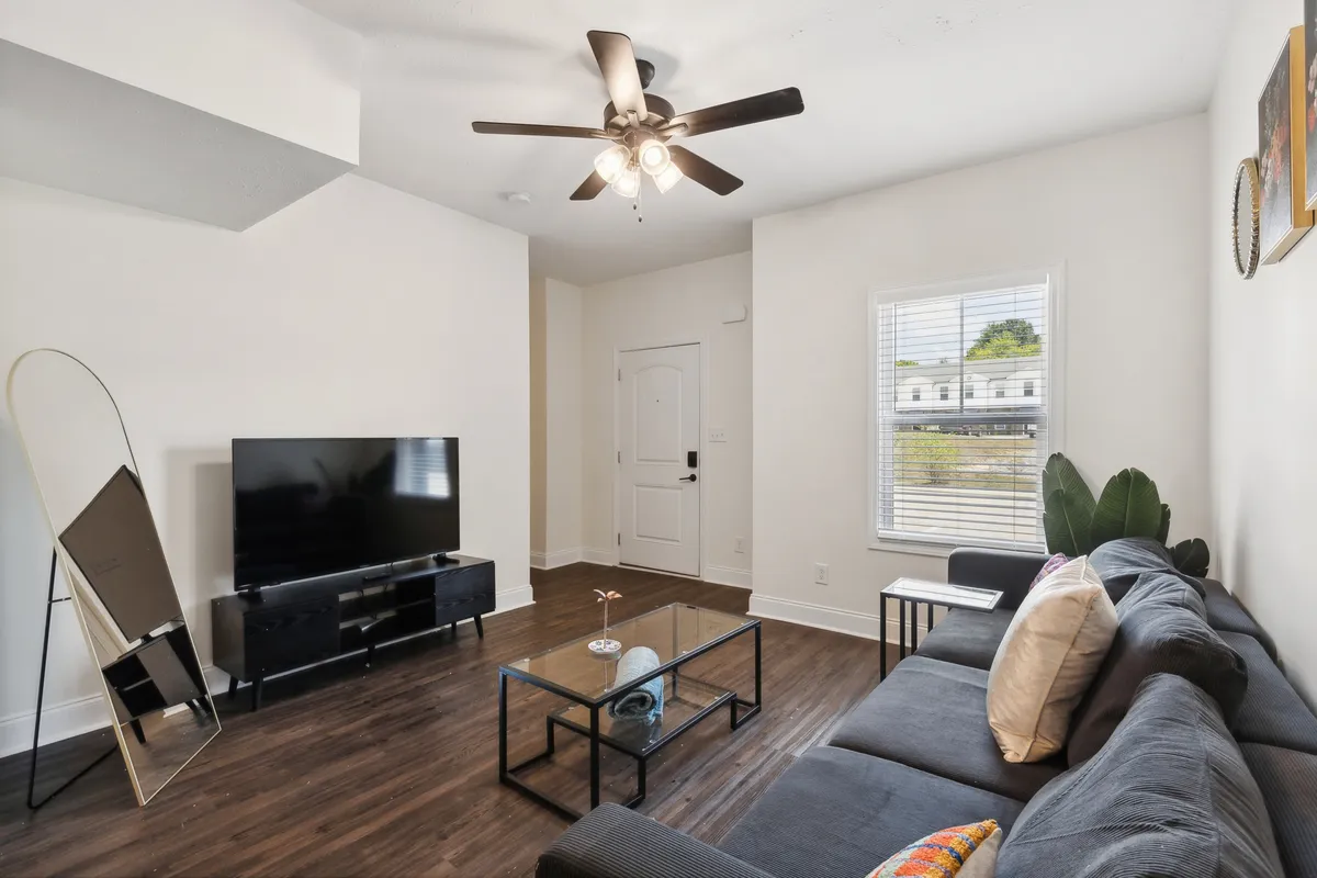 A cozy living room scene featuring a plush sofa, coffee table, television, and decorative mirror. The space exudes comfort and modern style, with sunlight streaming through windows, inviting guests to relax, socialize, or enjoy movie nights.